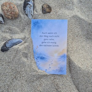 Puede incluir: Una tarjeta rectangular con una ilustración en acuarela azul y naranja de un camino que conduce hacia un amanecer, con el texto en alemán: Auch wenn ich den Weg noch nicht ganz sehe, gehe ich mutig den nächsten Schritt. La tarjeta está colocada en la arena.