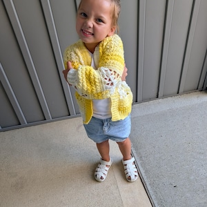 Può includere: Un cardigan all'uncinetto giallo e bianco con maniche lunghe e apertura frontale, indossato sopra una canotta bianca semplice, abbinato a pantaloncini di jeans azzurri e sandali bianchi con cinturini, creando un outfit a strati strutturato.