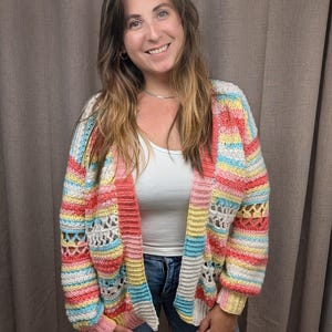 May include: A long-sleeve crochet cardigan featuring horizontal stripes in pastel shades of pink, yellow, blue, and white. The garment includes openwork stitch patterns on the sleeves and body, with ribbed cuffs and a ribbed front opening.