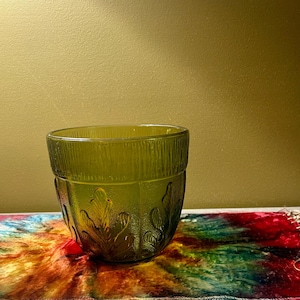 May include: A translucent olive green glass planter featuring a textured vertical ribbed pattern around the upper rim and a raised embossed leaf design encircling the lower body. The vessel has a wide circular opening and a rounded base.
