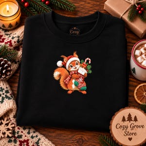 May include: A black crewneck sweatshirt featuring a central graphic of a cartoon squirrel wearing a red Santa hat and a matching scarf. The squirrel holds a red and white Christmas stocking filled with a white beverage and a candy cane.