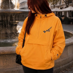 May include: A bright orange pullover windbreaker jacket featuring a half-zip closure, a drawstring hood, and a front kangaroo pocket. The chest displays an embroidered graphic of three small blue birds in flight, and a small logo patch on the sleeve.