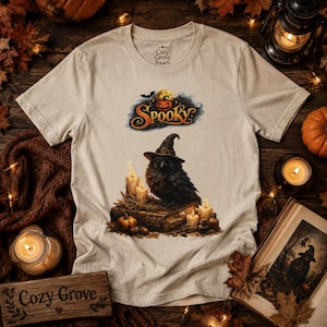 May include: A beige short-sleeve t-shirt featuring a graphic of a black crow wearing a witch hat perched on an old book. The design includes lit candles, a pumpkin, and the word "SPOOKY" in orange script. The brand name "Cozy Grove" is printed on the inner neck label.