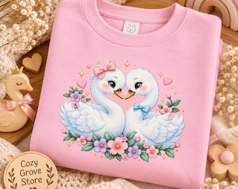 May include: A light pink crewneck sweatshirt featuring a graphic of two white swans facing each other, surrounded by pink and purple flowers, small hearts, and sparkles. The left swan wears a pink bow, and the right swan wears a blue bow.