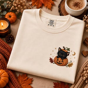 Könnte beinhalten: Ein cremefarbenes Sweatshirt mit Rundhalsausschnitt und einer kleinen Grafik auf der linken Brust. Das Motiv zeigt ein schwarzes Kätzchen, das in einem geschnitzten orangefarbenen Kürbis sitzt, umgeben von Herbstblättern, kleinen Sternen und einer Mondsichel.