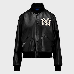 May include: A black leather varsity jacket featuring a white New York Yankees logo on the left chest, a snap-button front closure, ribbed black cuffs and hem, and a small white embroidered signature detail on the lower right side of the front panel.