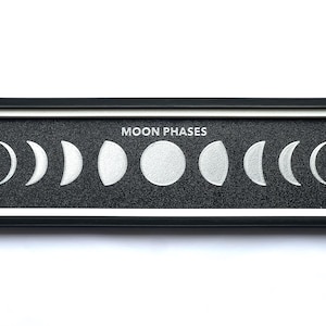 May include: A rectangular wall art piece in a black frame featuring a black textured background with the white text MOON PHASES centred above a horizontal sequence of eight silver moon phase illustrations ranging from a crescent to a full moon.