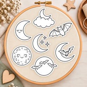 Pode incluir: Uma coleção de sete autocolantes a preto e branco dispostos num bastidor de bordar circular de madeira. Os desenhos apresentam uma lua crescente sorridente, uma lua atrás de uma nuvem, um morcego voador, estrelas e uma lua cheia.
