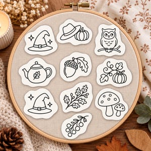 May include: A round wooden embroidery hoop containing beige fabric with ten black and white line art stickers featuring a witch hat, a hat with a pumpkin, an owl, a teapot, an acorn, a pumpkin vine, a second witch hat, an oak branch, mushrooms, and berries.