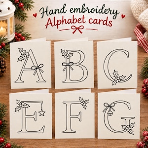 Puede incluir: Seis tarjetas plegadas de color blanco roto con letras A, B, C, E, F y G en arte lineal negro, decoradas con motivos navideños como acebo, bayas, lazos y estrellas. El texto "Hand embroidery Alphabet cards" aparece sobre las tarjetas.