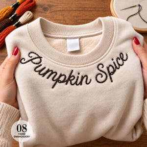 May include: A beige crewneck sweatshirt featuring the words Pumpkin Spice embroidered in dark brown cursive thread across the chest. The garment has a ribbed collar and a soft fleece interior lining, with a blank white label at the neckline.