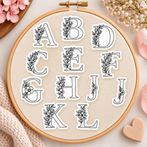 Può includere: Una collezione di adesivi in vinile bianco con le lettere dalla A alla L in un carattere a contorno nero in grassetto. Ogni lettera è decorata con un delicato disegno a linee nere di un tralcio floreale. Gli adesivi sono disposti su una superficie circolare in tessuto.