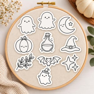 May include: A collection of nine black and white vinyl stickers arranged on a circular wooden embroidery hoop. The designs include ghosts, a crescent moon, a pumpkin, a potion bottle, a witch hat, a leafy branch, a bat, and sparkling stars.