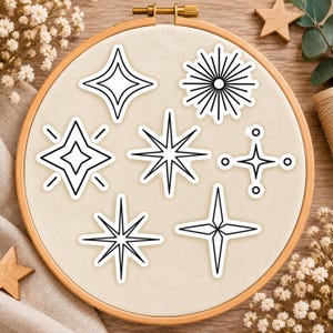 Puede incluir: Un bastidor de bordado de madera que contiene un fondo de tela de color crema con siete pegatinas o parches distintos en forma de estrella en blanco y negro, con varios diseños geométricos que incluyen rayos de sol y estrellas de cuatro puntas.