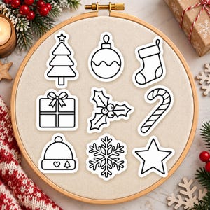 Könnte beinhalten: Ein hölzerner Stickrahmen mit einer beigen Stoffbespannung, auf der neun schwarz-weiße Aufkleber mit Umrissen von einem Weihnachtsbaum, einer Kugel, einem Strumpf, einem Geschenk, Stechpalmen, einer Zuckerstange, einer Mütze, einer Schneeflocke und einem Stern zu sehen sind.