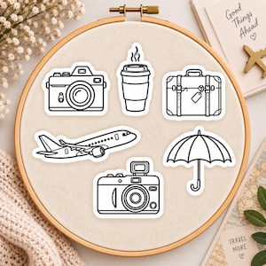 May include: A set of five black and white line art stickers featuring a camera, a coffee cup, a suitcase, an aeroplane, and an umbrella. The stickers are arranged on a circular embroidery hoop frame with a light fabric background.