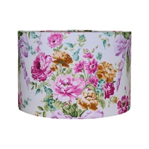 Puede incluir: Una pantalla de lámpara blanca con un patrón floral con flores rosas, amarillas y verdes. La pantalla de la lámpara tiene una forma cilíndrica y está hecha de tela.