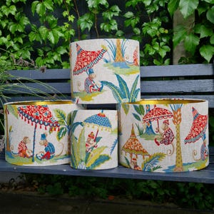 Puede incluir: Cuatro pantallas de lámpara cilíndricas con un borde dorado y un fondo blanco. Las pantallas de lámpara presentan un patrón colorido de monos, palmeras y paraguas.