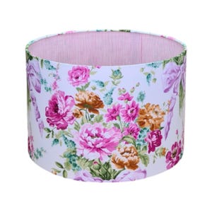 Puede incluir: Una pantalla de lámpara blanca con un patrón floral con flores rosas, naranjas y verdes. La pantalla tiene un forro a rayas rosa y blanco.