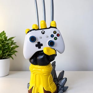 May include: A yellow and black gauntlet-style controller stand featuring three long silver claws extending upwards. The stand holds a white Xbox wireless controller with blue thumbsticks, a black directional pad, and multicoloured A, B, X, and Y buttons.