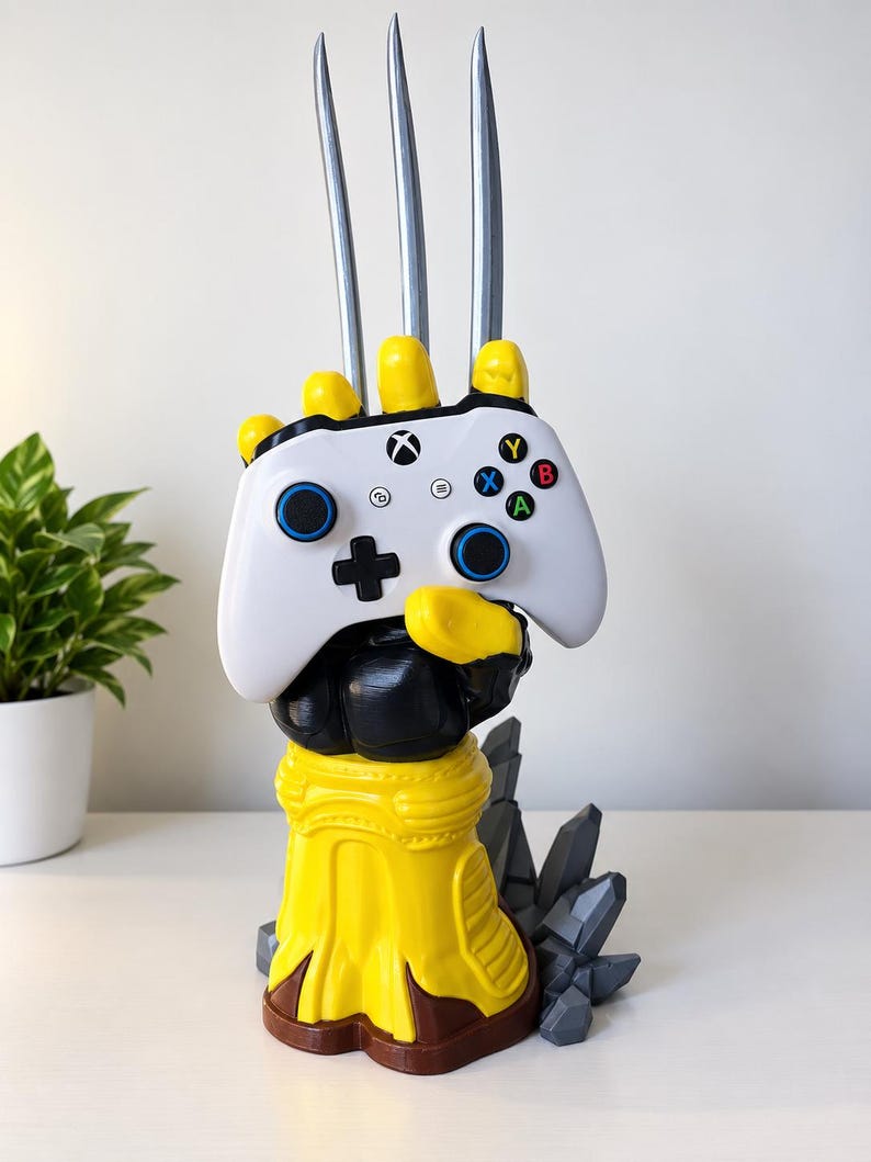 May include: A yellow and black gauntlet-style controller stand featuring three long silver claws extending upwards. The stand holds a white Xbox wireless controller with blue thumbsticks, a black directional pad, and multicoloured A, B, X, and Y buttons.