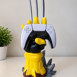 May include: A yellow and black hand-shaped controller stand featuring three long silver metallic claws extending upwards from the knuckles. The stand holds a white and black wireless game controller with standard directional buttons and analogue sticks.