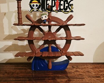 May include: A brown ship wheel display shelf with three horizontal tiers, a blue wave base, and a One Piece logo sign at the top. A small black flag with a white skull and crossbones emblem is attached to a mast on the left side of the structure.