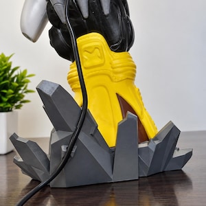 Pode incluir: Uma base de figura com uma bota amarela com um emblema quadrado, sobre uma formação rochosa geométrica cinzento-escura. Um cabo preto pende à frente da bota amarela e elementos prateados em forma de garra sobressaem do topo.