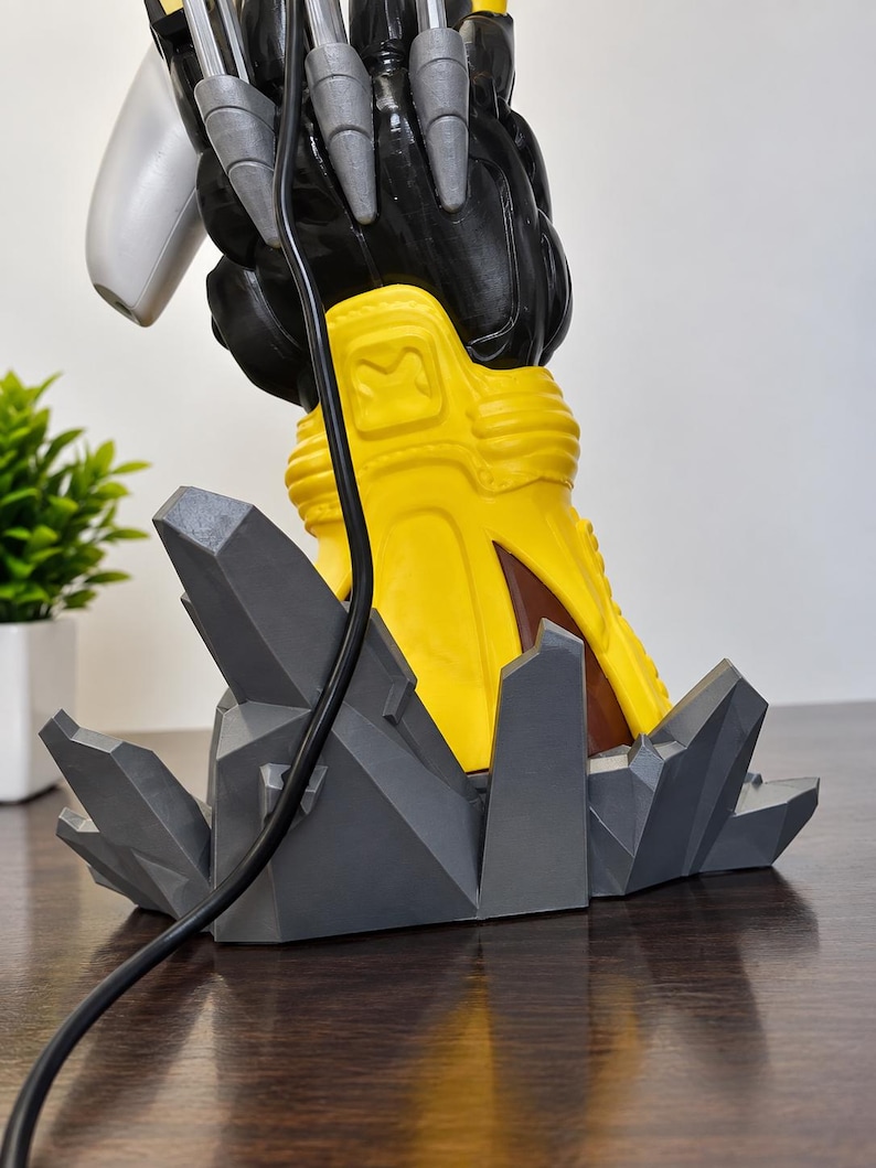 May include: A figurine base featuring a yellow boot with a square emblem, standing on a jagged, dark grey, geometric rock formation. A black cable hangs down the front of the yellow boot, and silver claw-like elements extend from the top.