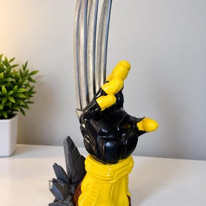May include: A decorative sculpture featuring a yellow and black gloved hand with three long, silver-coloured metallic blades extending upwards. The base is brown and yellow with grey, angular, rock-like formations attached to the side of the structure.