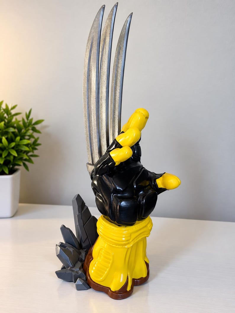May include: A decorative sculpture featuring a yellow and black gloved hand with three long, silver-coloured metallic blades extending upwards. The base is brown and yellow with grey, angular, rock-like formations attached to the side of the structure.
