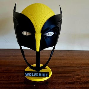 Peut inclure: Un masque jaune et noir avec une surface texturée, des protubérances pointues en forme d'oreilles et des caches oculaires en maille blanche. Le masque est monté sur un support noir avec une base circulaire jaune portant le mot WOLVERINE.
