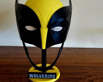 May include: A yellow and black mask featuring a textured surface, pointed ear-like protrusions, and white mesh eye coverings. The mask is mounted on a black stand with a yellow circular base that displays the word WOLVERINE in white lettering.