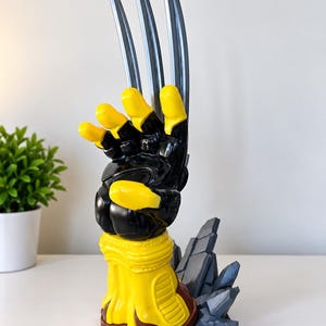 May include: A sculpted hand figurine featuring a glossy black and yellow colour scheme with three long, silver-coloured metallic blades extending from the knuckles. The base is yellow with brown accents, supported by a cluster of jagged, grey rock-like structures.