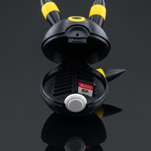 Può includere: Un contenitore sferico nero e giallo a forma di creatura con orecchie a punta. Il contenitore è aperto e mostra fessure interne per cartucce di gioco, con una scheda di gioco Nintendo Switch inserita. Sulla parte anteriore c'è un bottone bianco.