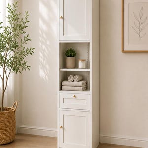 May include: A tall, white, narrow storage cabinet featuring a top cabinet door, an open middle section with two shelves, a single drawer, and a bottom cabinet door. Each door and the drawer are equipped with a small, round, gold-colored knob.