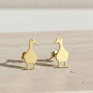 May include: A pair of gold-colored duck-shaped stud earrings. The earrings are simple in design, with a smooth, polished finish. The ducks are standing upright, with their beaks pointing upwards. The earrings are set against a neutral background.