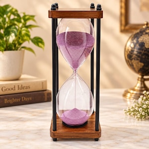 Puede incluir: Un reloj de arena con marco de madera y cuatro pilares de metal negro. El bulbo de vidrio transparente contiene arena fina de color púrpura, con una parte fluyendo actualmente por el centro hacia la cámara inferior. La base y la parte superior son de madera pulida.