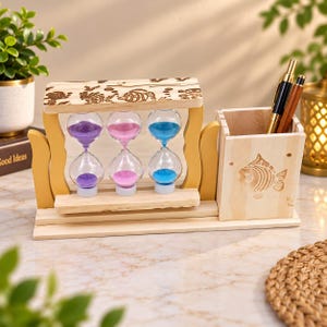 May include: A wooden desk organiser featuring three hourglasses with purple, pink, and blue sand. The frame has engraved sea life patterns, and an attached square pen holder displays an engraved fish design. Two pens are stored inside the holder.