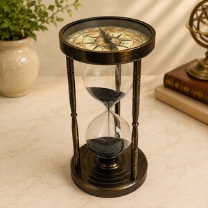 May include: An antique-style hourglass with a dark bronze metal frame and black sand. The top of the frame features a circular compass with a glass cover, displaying cardinal directions and a central needle, supported by three vertical pillars.