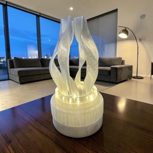 May include: A white, translucent, sculptural lamp featuring a cylindrical base and a series of upward-curving, flame-like vertical fins. A string of warm-toned LED lights is coiled around the base, illuminating the textured, semi-transparent structure.