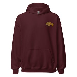 May include: A maroon hooded sweatshirt featuring a small embroidered graphic on the left chest. The embroidery depicts a stylized bison in shades of gold, brown, and cream with geometric patterns integrated into the animal's body design.