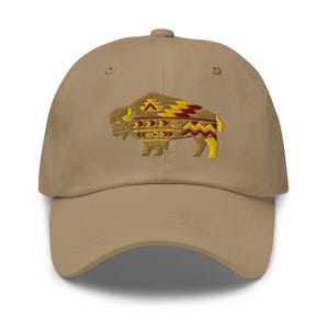 Puede incluir: Una gorra de béisbol de color caqui con un gráfico bordado de un bisonte en la parte delantera. El bisonte está relleno de patrones geométricos en tonos amarillo, rojo oscuro y marrón, creando un aspecto detallado y texturizado.