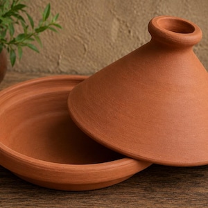 May include: A terracotta tagine cooking vessel consisting of a wide, shallow circular base and a tall, conical lid with a small opening at the top. The unglazed clay surface has a matte, earthy reddish-brown color and a smooth, uniform texture.
