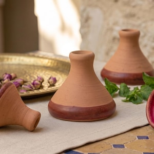 May include: A collection of terracotta clay bowls with a wide base and narrow neck. The exterior is a natural matte tan color with a dark reddish-brown glazed band at the base. The interior of the bowls features a vibrant, glossy magenta finish.