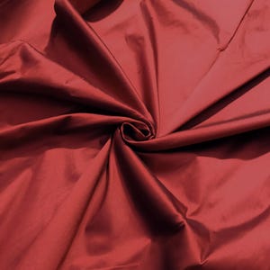Puede incluir: Una tela de color rojo intenso con una superficie lisa y ligeramente brillante. El material está recogido en el centro, creando pliegues radiantes y sombras suaves que resaltan la textura y el rico color carmesí sólido del tejido.