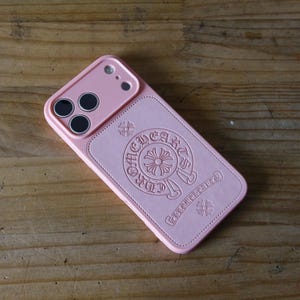 May include: A light pink smartphone case featuring a raised camera cutout and a central embossed circular logo design with the text CHROME HEARTS. The case has a smooth, matte finish and a rectangular shape with rounded corners and stitched edges.