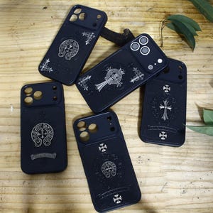 Op de afbeelding: Vier zwarte smartphonehoesjes met zilverkleurige metalen emblemen en ingewikkelde kruismotieven. Elk hoesje heeft een rechthoekige uitsparing voor de camera en een gladde, matte afwerking met cirkelvormige logo's en decoratieve accenten.