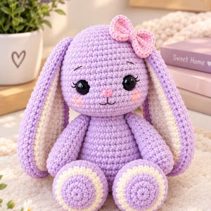 May include: A lavender crochet bunny plush toy featuring long ears with cream-colored inner panels, large black safety eyes, a small pink stitched nose, and a light pink bow attached to the top of its head. The bunny has round feet with cream accents.