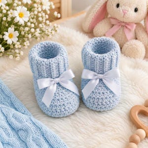 May include: A pair of light blue knitted baby booties featuring a ribbed cuff and a white satin ribbon bow tied at the front. The booties are crafted with a textured stitch pattern and are presented alongside a plush cream-colored rabbit toy.